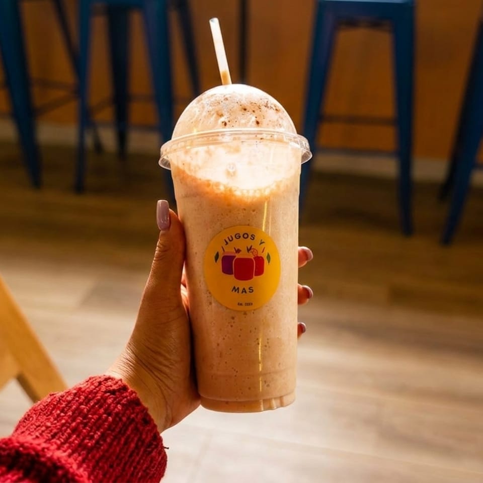 Hand holding a Jugos y Mas smoothie with a straw, against a blurred background of blue bar stools.