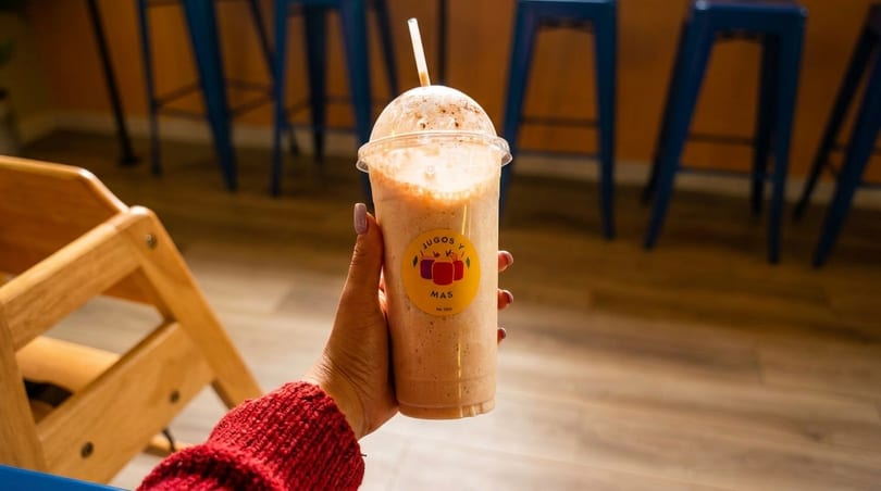 Hand holding a Jugos y Mas smoothie with a straw, against a blurred background of blue bar stools.