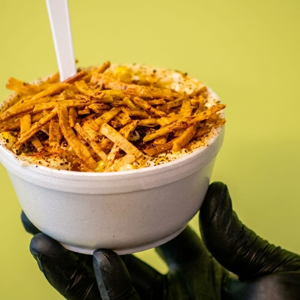 A hand in a black glove holds a white cup of elote topped with crispy tortilla strips.