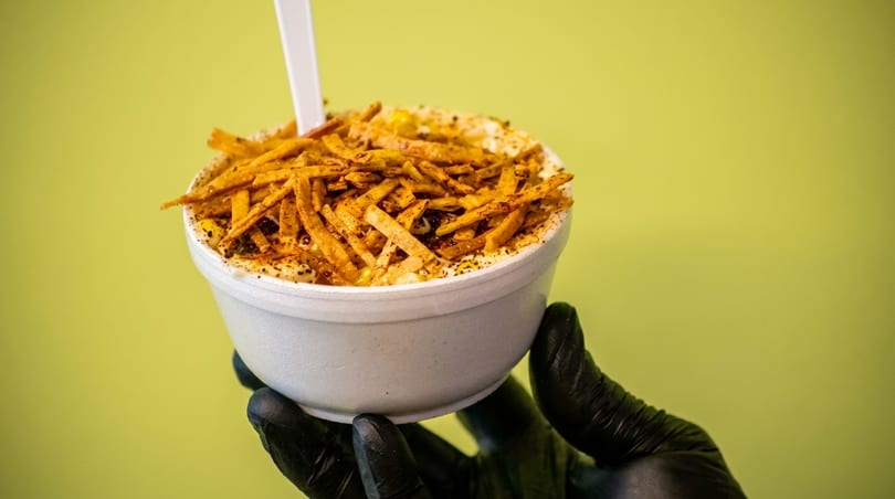 A hand in a black glove holds a white cup of elote topped with crispy tortilla strips.