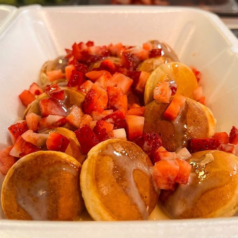 Mini pancakes topped with diced strawberries and a sweet glaze in a white takeout container.