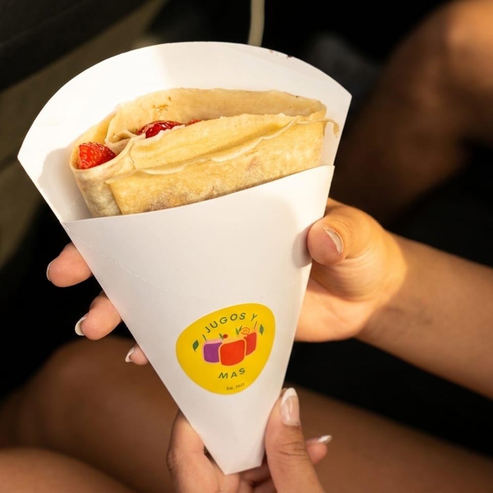A person holds a crepe filled with strawberries and cream in a white paper cone from Jugos y Mas.