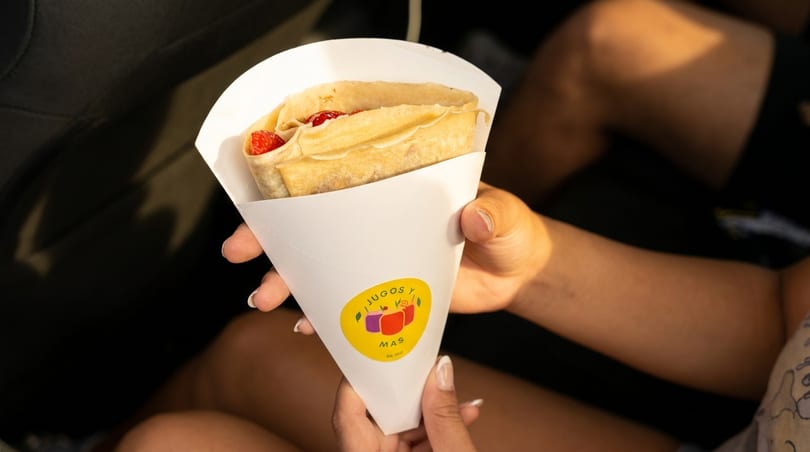 A person holds a crepe filled with strawberries and cream in a white paper cone from Jugos y Mas.